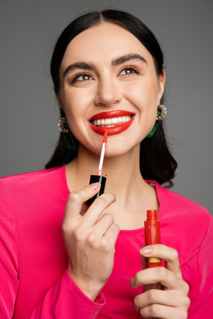 alluring young woman with shiny brunette hair and trendy earrings posing in magenta dress while applying red lip gloss on lips and smiling isolated on grey backgroundの写真素材