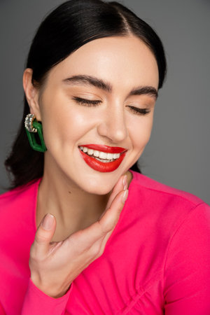 portrait of happy young woman with shiny brunette hair, trendy earrings and red lips smiling with closed eyes while posing isolated on grey backgroundの写真素材