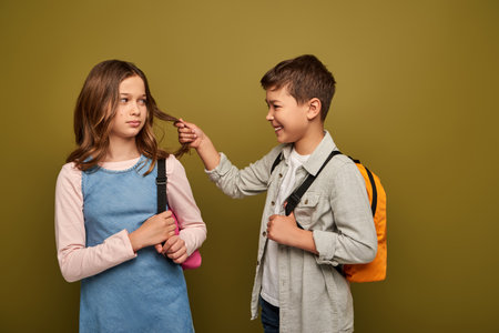 Smiling multiracial boy with backpack pulling hair of preteen girl while getting attention during international child protection day celebration on khaki backgroundの写真素材