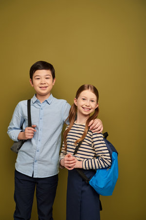 Smiling asian boy with backpack hugging red haired friend and looking at camera during international...