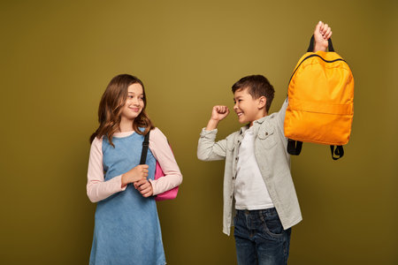 Excited multiracial boy holding backpack and showing yes gesture near friend in dress standing and smiling during child protection day celebration on khaki backgroundの写真素材