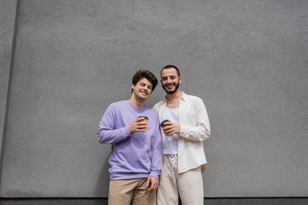 Smiling and young same sex couple in casual clothes holding coffee to go and looking at camera while standing near building outdoorsの写真素材