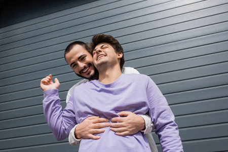 Low angle view of positive and bearded gay man hugging brunette boyfriend in sweatshirt and braces...