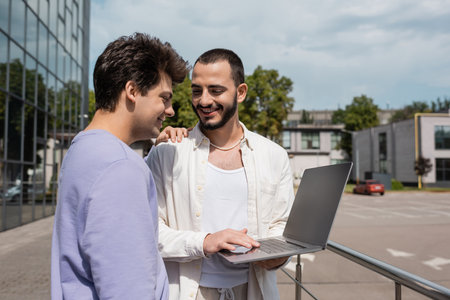Positive gay man in casual clothes hugging bearded boyfriend using laptop with blank screen near...