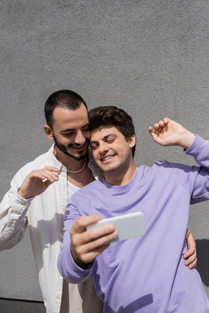 Overjoyed homosexual man in casual clothes using mobile phone while standing next to boyfriend in braces and building on urban street outdoorsの写真素材