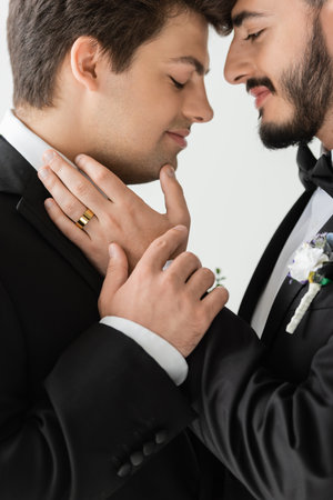 Side view of young same sex grooms in classic suits with boutonnieres touching each other during wedding celebration and while standing isolated on greyの写真素材