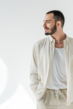 Bearded and short haired homosexual man in beige shirt and stylish pearl necklace looking away and...