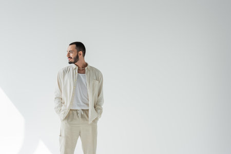 Smiling homosexual man in beige clothes and pearl necklace holding hands in pockets of pants and looking away while standing on grey background with sunlightの写真素材