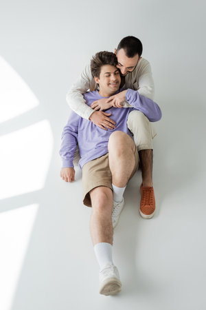 High angle view of brunette gay man in casual clothes embracing cheerful partner in braces with...