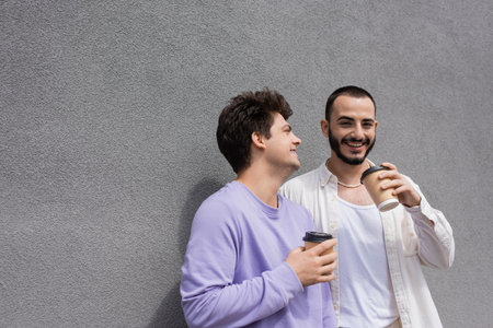 Happy bearded homosexual man holding coffee to go near boyfriend in purple sweatshirt and braces...