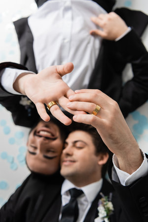 Top view of blurred same sex grooms in suits touching fingers of each other in wedding rings while lying on festive confetti on grey backgroundの写真素材