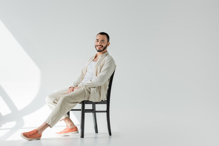 Positive brunette gay man in casual clothes made of natural fabrics looking at camera while sitting on comfortable chair on grey background with sunlightの写真素材