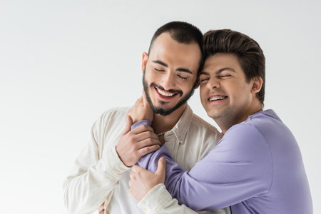 Carefree young gay man in braces wearing casual clothes hugging and touching neck of brunette and bearded partner with closed eyes isolated on greyの写真素材