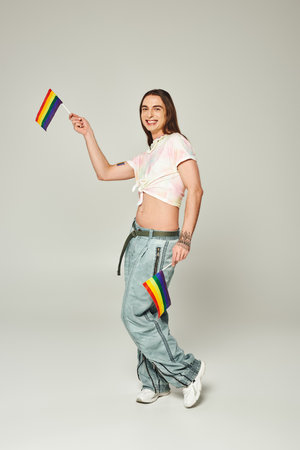 cheerful and tattooed gay man with long hair and bare belly standing in denim jeans while holding rainbow flags for pride month on grey backgroundの写真素材