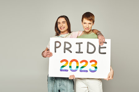 cheerful lgbt friends holding pride 2023 placard and looking at camera while celebrating lgbtq community holiday in June on grey background in studioの写真素材