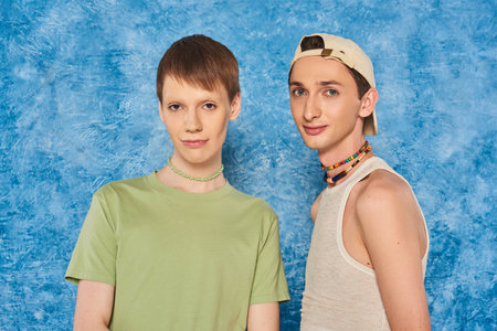young lgbtq friends with colorful beads and casual clothes and looking at camera while standing together on mottled blue background during pride monthの写真素材