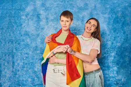 Smiling tattooed gay man in casual clothes hugging young friend with lgbt flag and looking at camera...