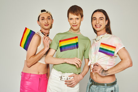 positive and young lgbt friends with tattoos standing in colorful clothes and holding rainbow flags while looking at camera in studio on pride monthの写真素材