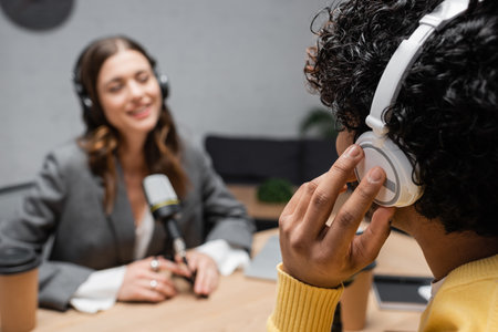 young indian man adjusting headphones during interview with blurred radio host in grey blazer smiling near microphone and paper cup on blurred background in broadcasting studioの写真素材