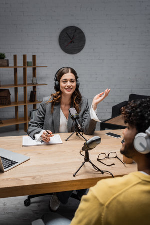Smiling podcast host in wireless headphones talking to blurred indian guest near notebook, pen, glasses, microphones and devices with copy space during stream in studioの写真素材