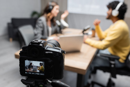 selective focus of professional digital camera recording how interviewer talking to young indian guest while sitting together near laptop and microphones on blurred background in studioの写真素材