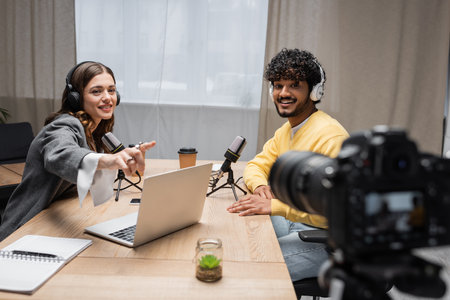 cheerful broadcaster in headphones pointing at laptop near coffee to go and notebook during...