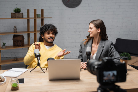 positive indian man in yellow jumper gesturing near microphones, laptop, notebook and smartphone while talking to smiling colleague sitting in blazer in front of blurred digital camera in studioの写真素材