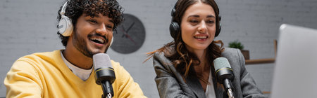 excited and curly indian man in yellow jumper with charming brunette woman in grey blazer talking in professional microphones while working and recording podcast in radio studio, bannerの写真素材