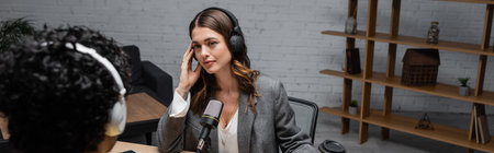 brunette radio host in grey blazer adjusting headphones during interview with blurred indian man near microphone and paper cup with wooden rack on background in broadcasting studio, bannerの写真素材
