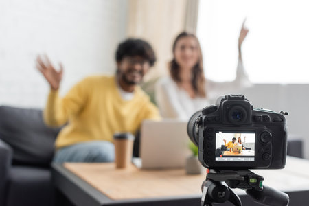 selective focus of professional digital camera near blurred interracial vloggers waving hands near...