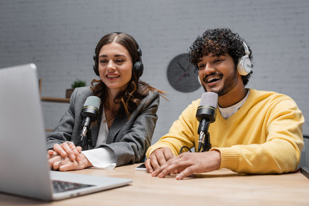 cheerful multiethnic radio hosts in headphones, yellow jumper and grey jacket looking at laptop...