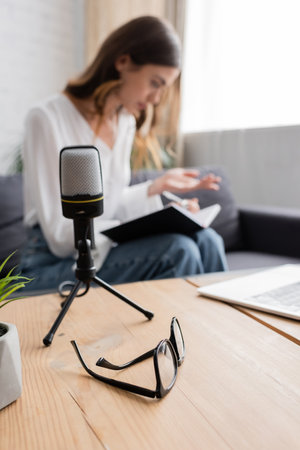 selective focus of table with laptop and eyeglasses and professional microphone near podcaster sitting on couch and working with notebook on blurred backgroundの写真素材