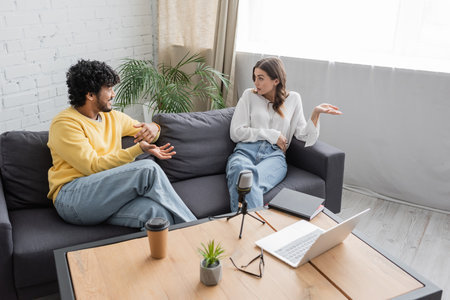 high angle view of multiethnic couple in casual clothes gesturing and talking on sofa near table with laptop, microphone, coffee to go, eyeglasses and flowerpot in radio studioの写真素材