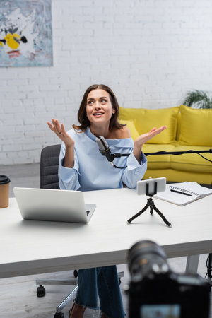 Smiling brunette podcaster looking away while talking near microphone, devices, coffee to go and...
