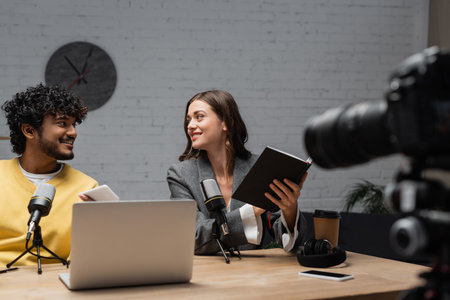 cheerful brunette podcaster in grey blazer sitting with notebook near young indian colleague holding smartphone near laptop, headphones and smartphone with blank screen in studioの写真素材
