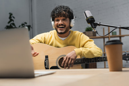 cheerful indian musician in headphones and yellow jumper playing acoustic guitar near blurred laptop and studio microphone on table with blurred coffee to go while recording podcast in studioの写真素材