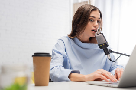 Brunette broadcaster talking at microphone and using blurred laptop near coffee to go in paper cup on table during stream in podcast studioの写真素材