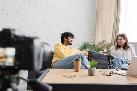 curly indian man in yellow jumper and brunette woman in white blouse talking on couch next to...