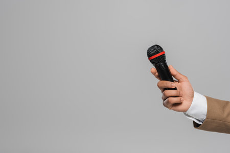 Cropped view of hand of event host in brown formal wear holding wireless microphone while standing isolated on grey with copy spaceの写真素材