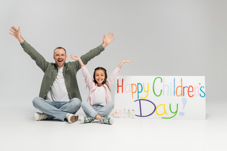 Excited and cheerful man and preteen daughter in casual clothes looking at camera together while sitting near placard with happy children's day lettering on grey backgroundの写真素材