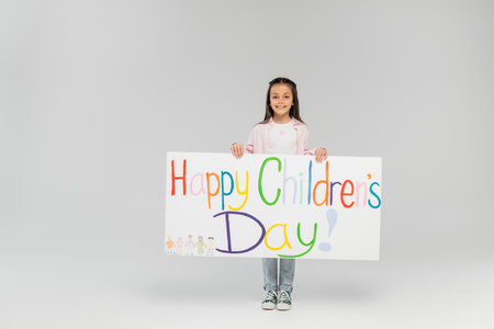 Full length of cheerful preteen girl in casual clothes looking at camera while holding placard with happy children's day lettering while standing on grey background with copy spaceの写真素材