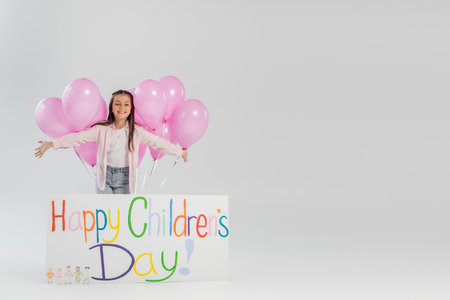 Cheerful and preteen girl in casual clothes looking at camera near pink festive balloons and placard with happy children's day lettering while celebrating on grey backgroundの写真素材