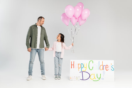 Full length of smiling preteen girl in casual clothes holding festive balloons and hand of father near placard with happy children's day lettering on grey backgroundの写真素材