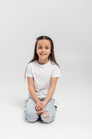 joyful preteen girl with long brunette hair sitting in white t-shirt and blue denim jeans and smiling isolated on grey background, child protection day holidayの写真素材