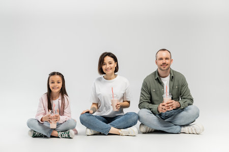 Smiling adult parents in casual clothes looking at camera while holding plastic cups with milkshakes near preteen daughter during international children day celebration and sitting on grey backgroundの写真素材