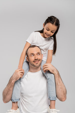 happy preteen daughter with long brunette hair sitting on shoulders of unshaved and cheerful father with bristle on grey background, Child protection day, father and daughterの写真素材
