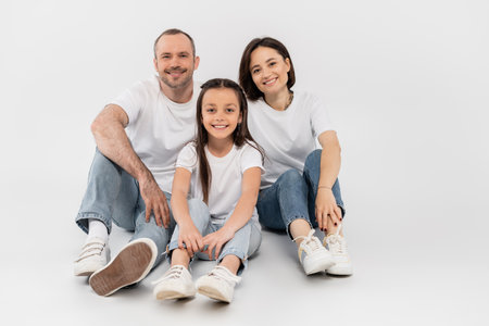stylish family in white t-shirts and blue denim jeans looking at camera and sitting together on grey background, International child protection day, happy parents and daughterの写真素材