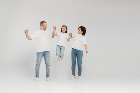 full length of happy parents in white t-shirts and blue denim jeans holding hands and lifting...