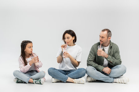 Adult parents in casual clothes drinking milkshakes from plastic cups with drinking straws and looking at preteen daughter during child protection day celebration while sitting on grey backgroundの写真素材