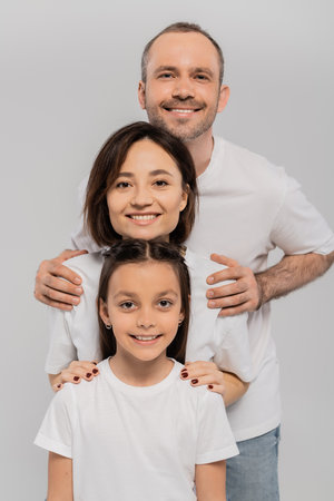 happy family in white t-shirts looking at camera and posing together on grey background, International child protection day, positive parents and daughterの写真素材